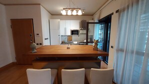A dining area with a wooden table and four chairs, a kitchen in the background, and a view through a window with curtains.