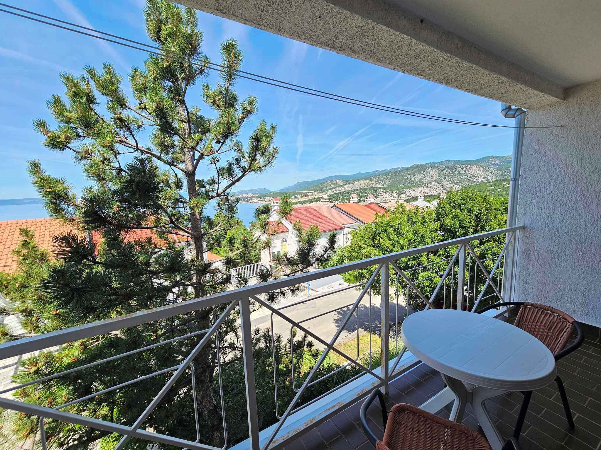 A balcony with a view of a coastal town, a table, and two wicker chairs.