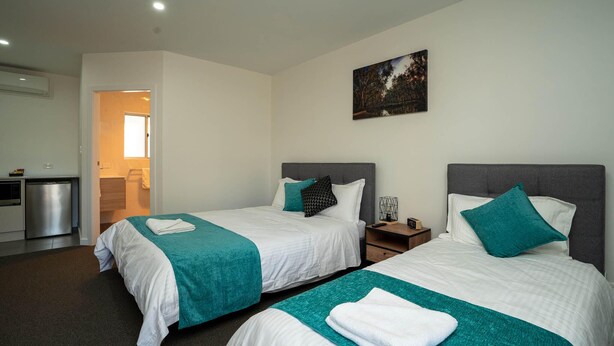 A hotel room with two beds, each with a white and teal bedspread, a grey headboard, and a teal pillow. There is a small bedside table with a lamp, a wall-mounted painting, and a door leading to a bathroom. - Brolga Hotel Motel (Coleambally)