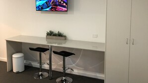 A hotel room with a TV mounted on the wall, a white desk with two bar stools, a small trash bin, and a wardrobe. - Brolga Hotel Motel (Coleambally)