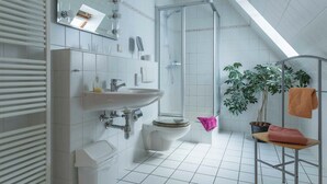 Moderni kylpyhuone, jossa on valkoinen pesuallas, wc ja suihku. Siellä on myös ruukussa oleva kasvi ja pyyheteline, jossa on pyyhkeitä.