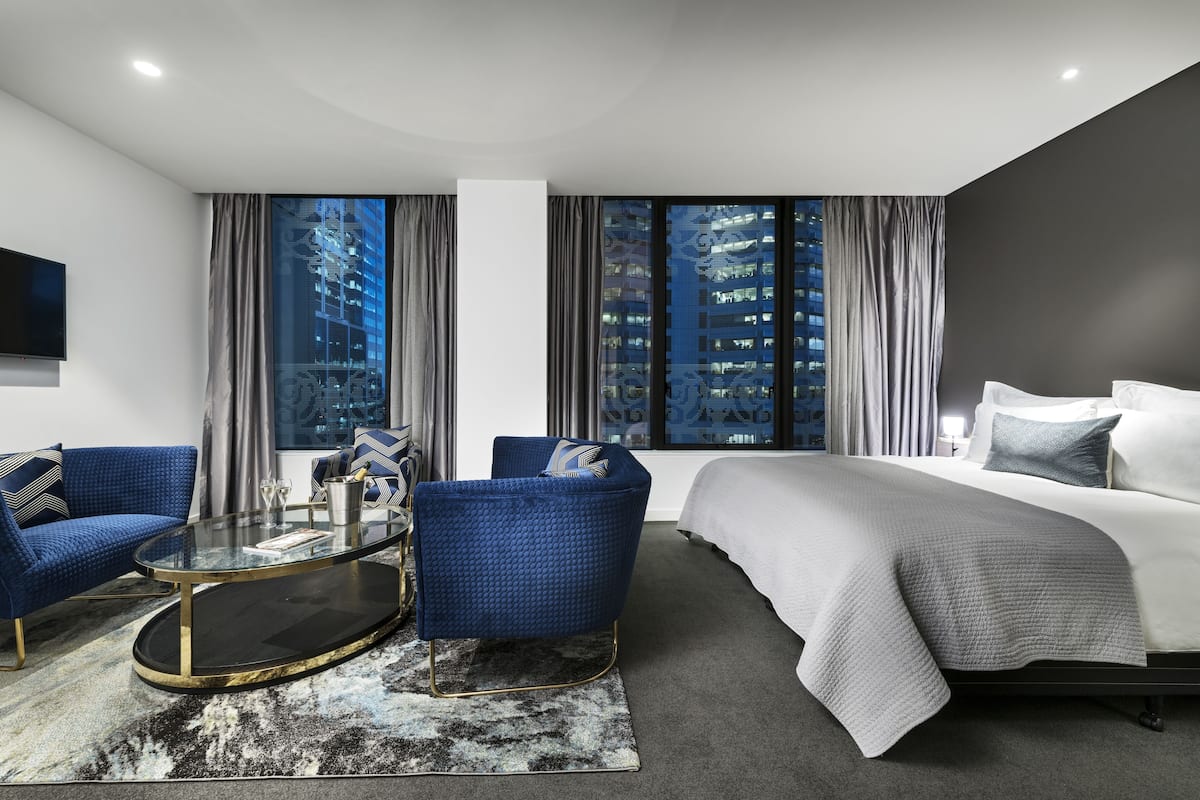 A modern hotel room with a large bed, a seating area with a glass coffee table, and a view of a cityscape through large windows.