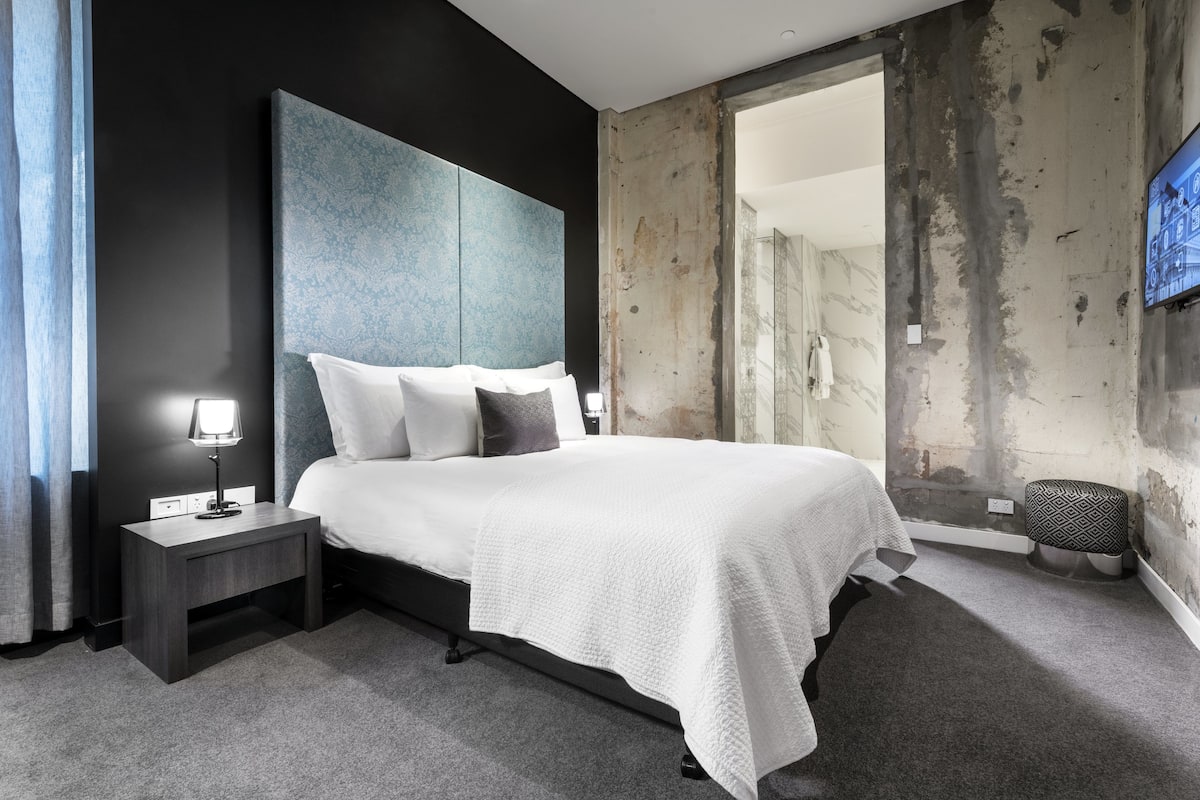 A modern hotel room with a large bed, a nightstand with a lamp, and a view through a glass door to a bathroom.