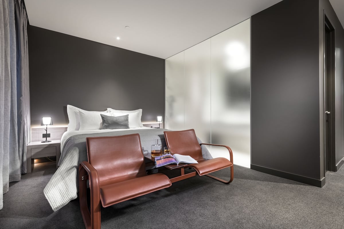 A modern hotel room with a large bed, two leather chairs, a bedside table with a lamp, and a large mirror wall.