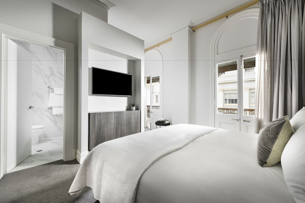 A modern hotel room with a large bed, a flat-screen TV mounted on the wall, a bathroom with a marble floor, and a view of the city through the window.