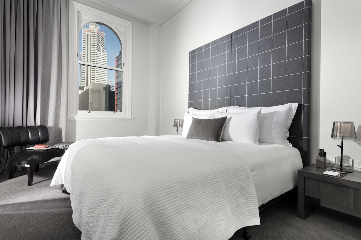 A modern hotel room with a large bed, a view of a cityscape through a window, a black leather chair, and a nightstand with a lamp.