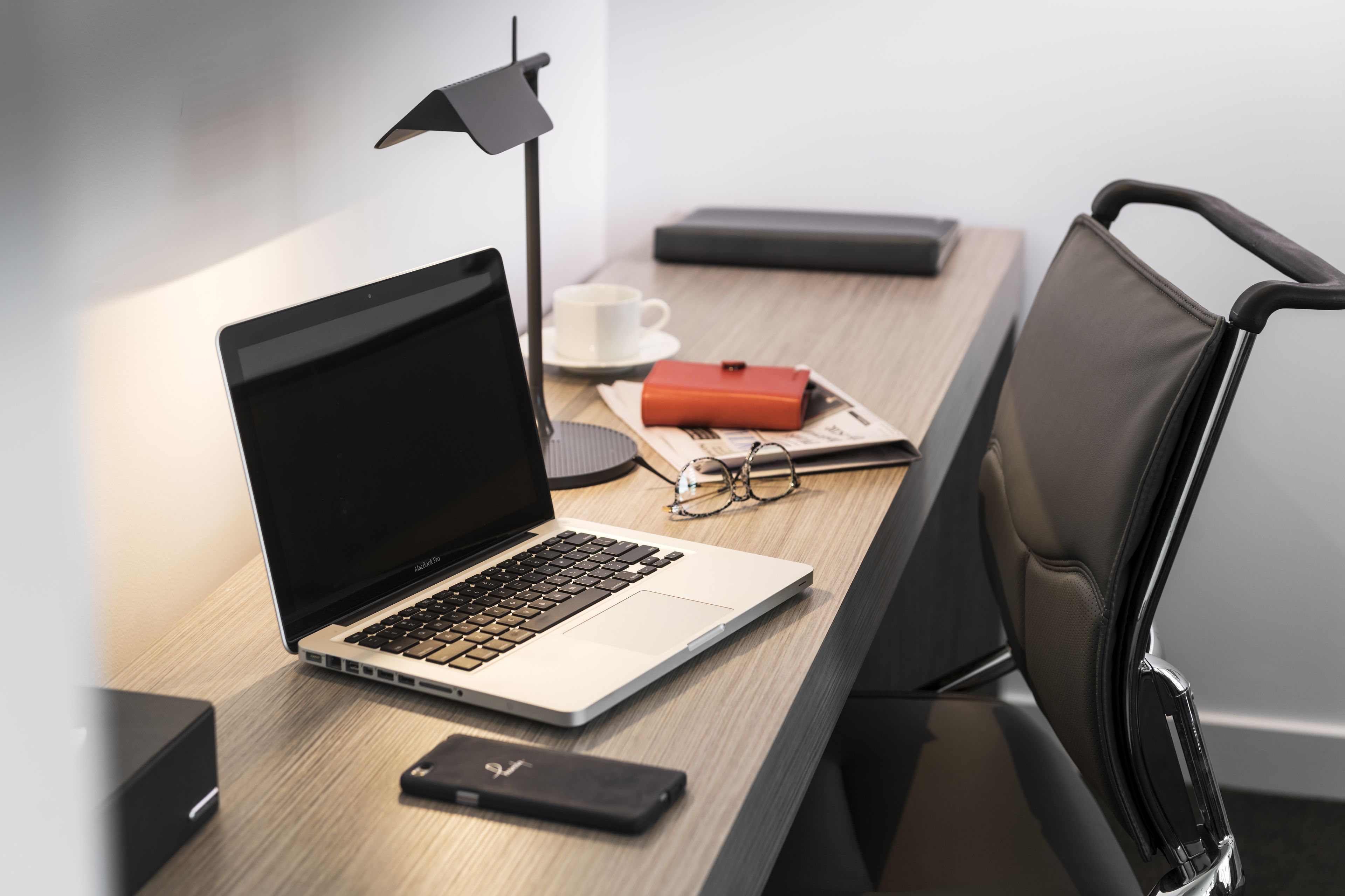 A laptop on a desk with a lamp, glasses, a cup, and a notebook.