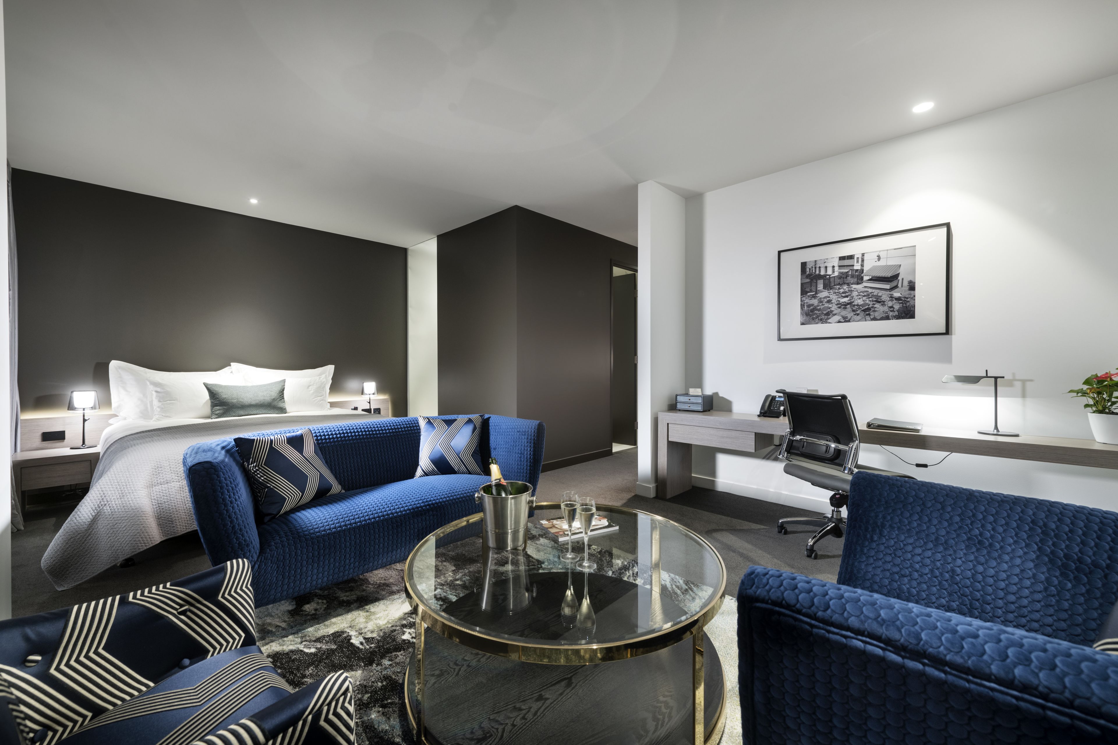 A modern hotel room with a bed, two blue sofas, a glass coffee table, a desk, and a framed picture on the wall.