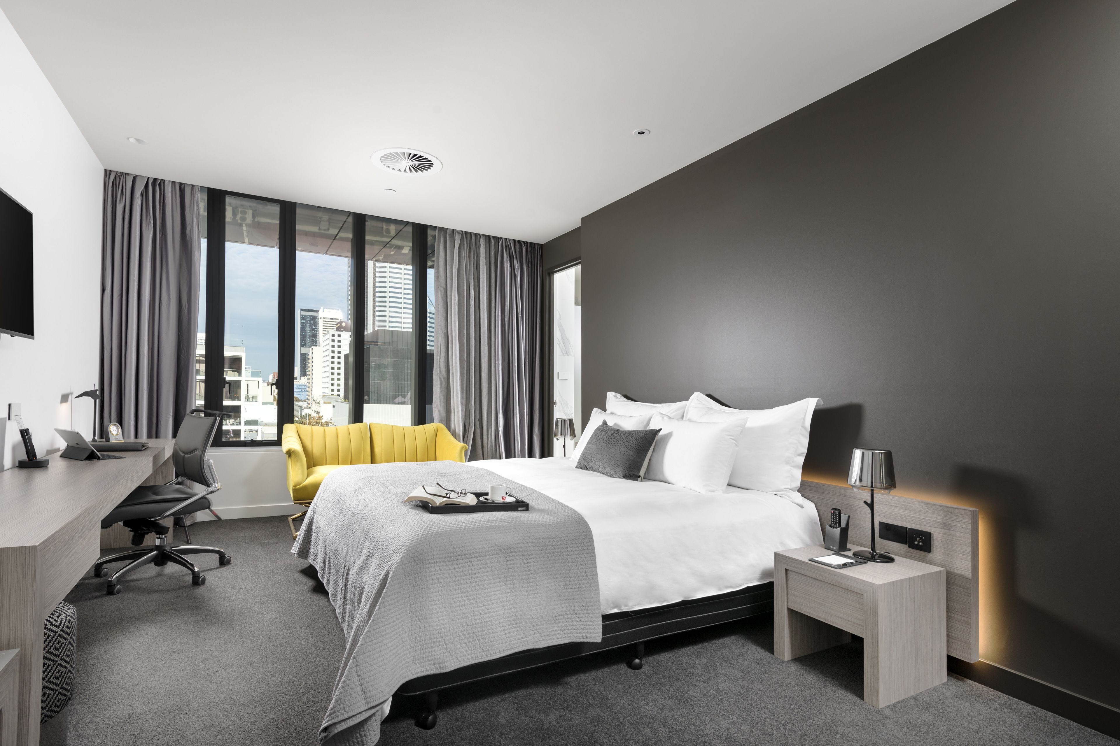 Ett modernt hotellrum med en stor säng, ett skrivbord, en stol och utsikt över staden genom stora fönster.