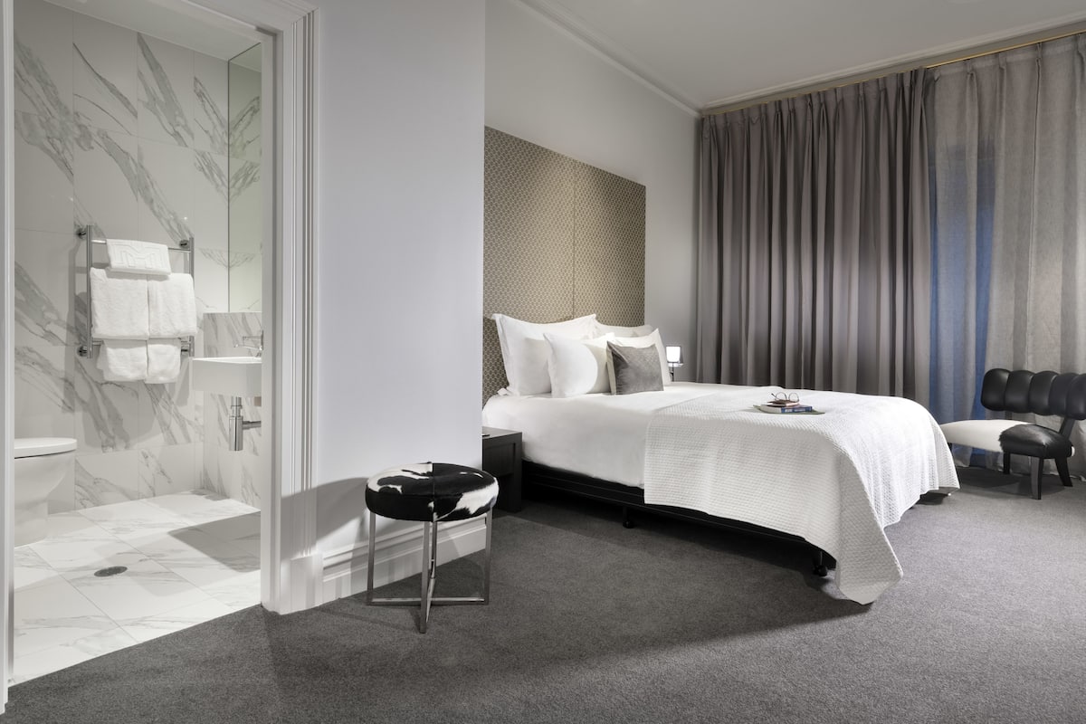 A modern hotel room with a marble bathroom, a large bed with white linens, a black leather chair, and a view of the city.