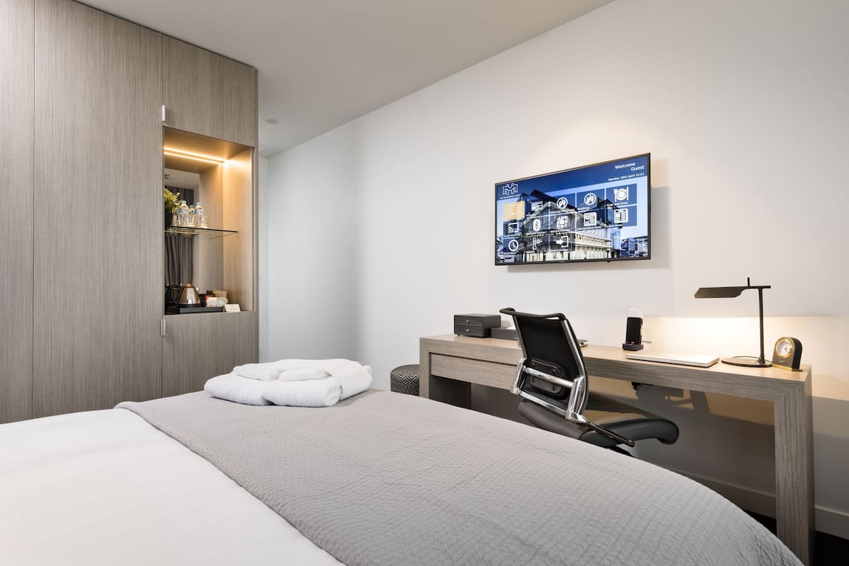 A modern hotel room with a bed, a desk, a chair, a desk lamp, a clock, and a framed picture on the wall.
