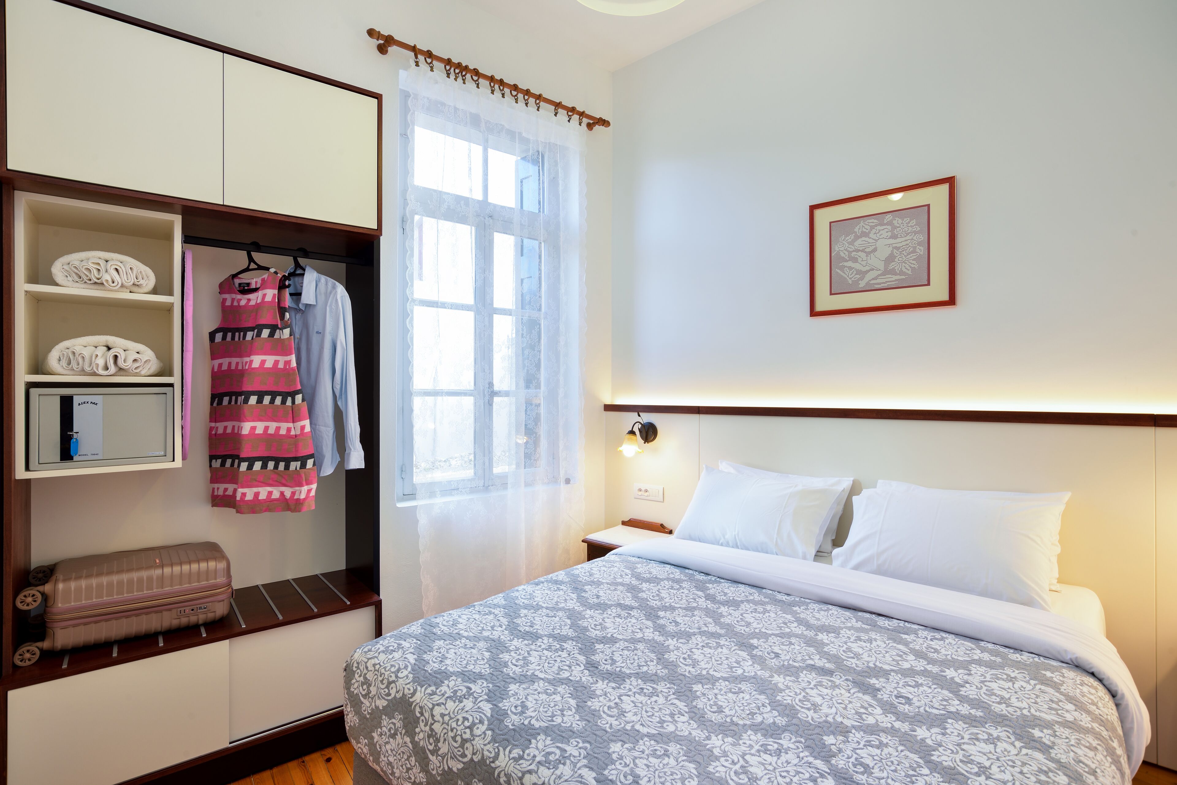 Een hotelkamer met een bed, een kledingkast, een raam met dunne gordijnen, een nachtkastje met een lamp, en een lijst met een afbeelding aan de muur.
