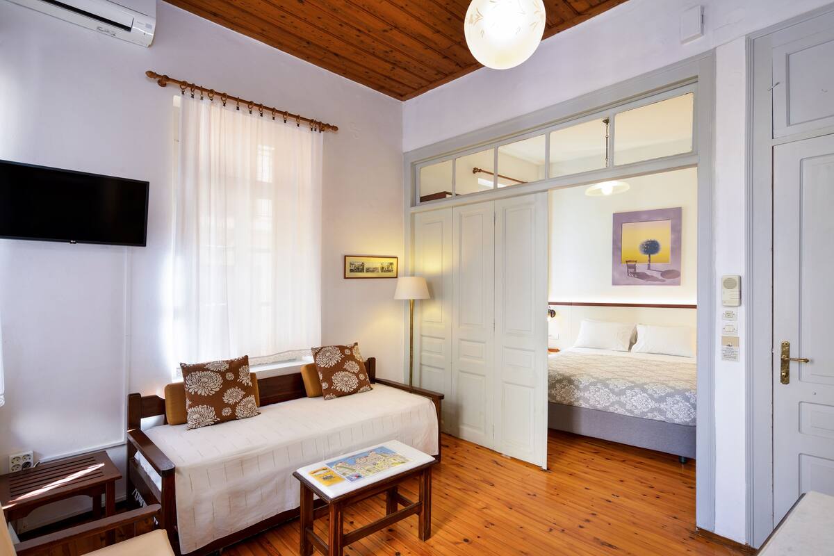 Quarto com piso de madeira, cama com colcha branca, mesa de cabeceira com um livro, janela com cortinas leves e TV fixada na parede.