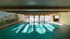 An indoor swimming pool with clear glass walls, wooden ceiling beams, and a mural on the left wall.