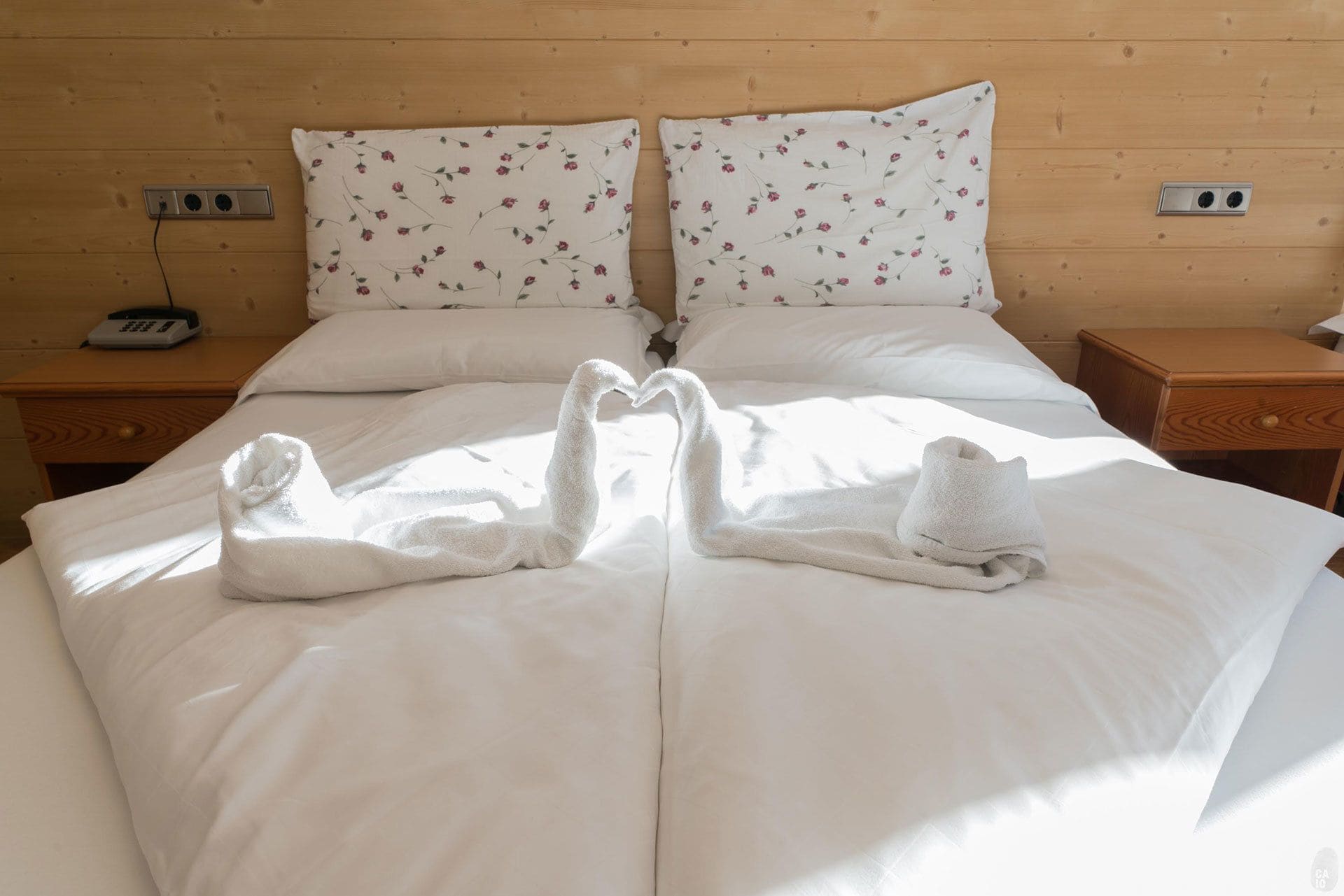 A neatly made bed with white linens and floral-patterned pillows, two towels arranged in a heart shape, and a bedside table with a telephone.