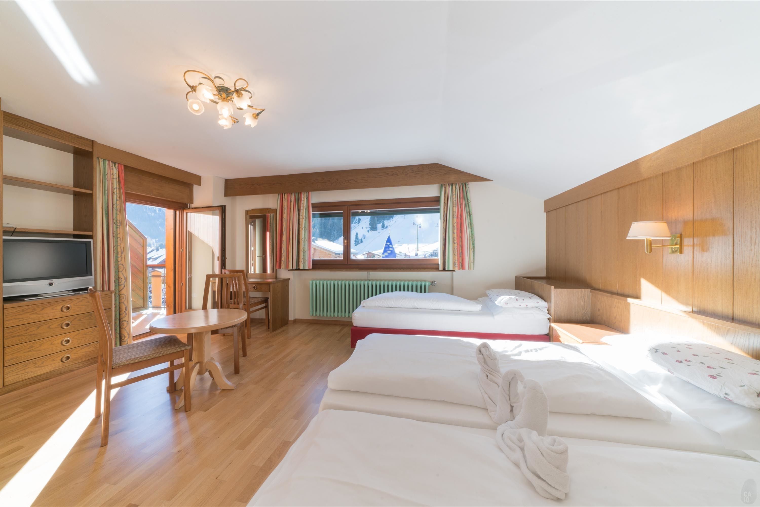 A hotel room with a wooden floor, a bed, a TV, a dining table with chairs, a window with curtains, and a lamp.