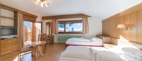 A hotel room with a wooden floor, a bed, a TV, a dining table with chairs, a window with curtains, and a lamp.