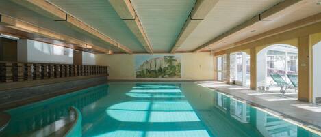 An indoor swimming pool with wooden beams on the ceiling, a mural on the wall, and a view of the outside through large windows.