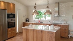 Une cuisine moderne avec des meubles en bois, des appareils électroménagers en acier inoxydable, un îlot central, des luminaires suspendus et une fenêtre donnant sur un espace verdoyant.