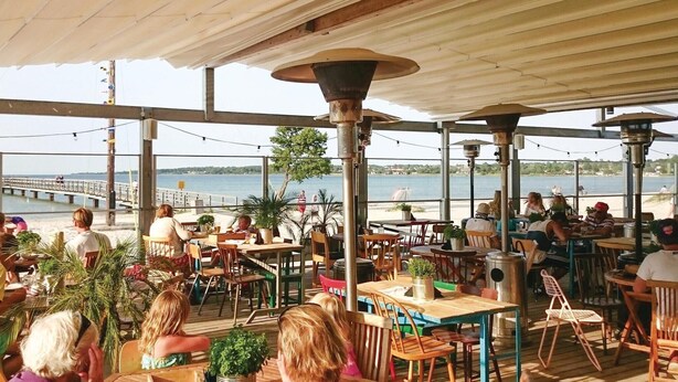 Un espace de restauration en plein air, avec des tables et des chaises, offrant une vue sur un plan d’eau et une jetée.