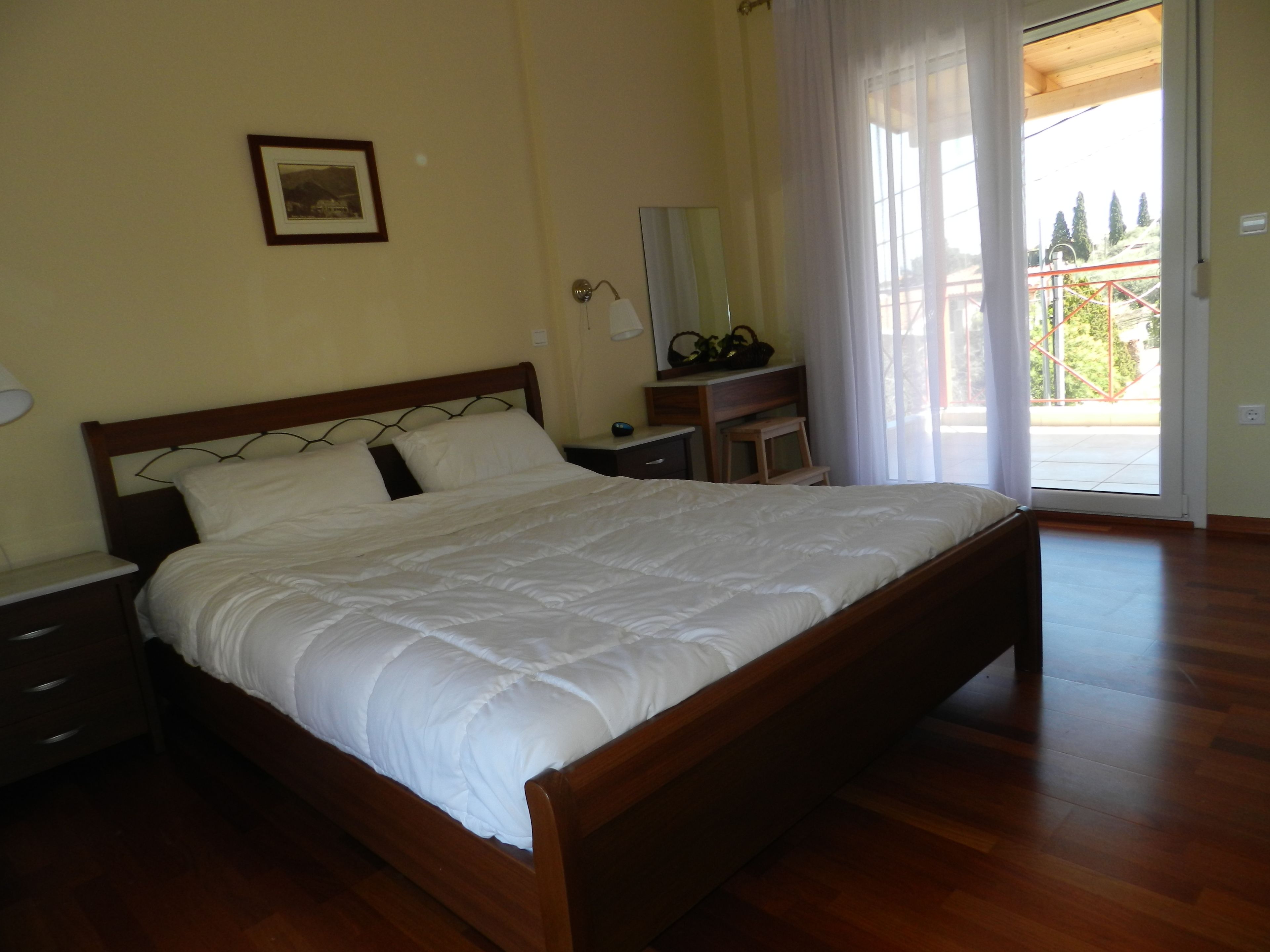 A bedroom with a wooden bed, a nightstand, a dresser, a mirror, and a window with a view of trees and a balcony.