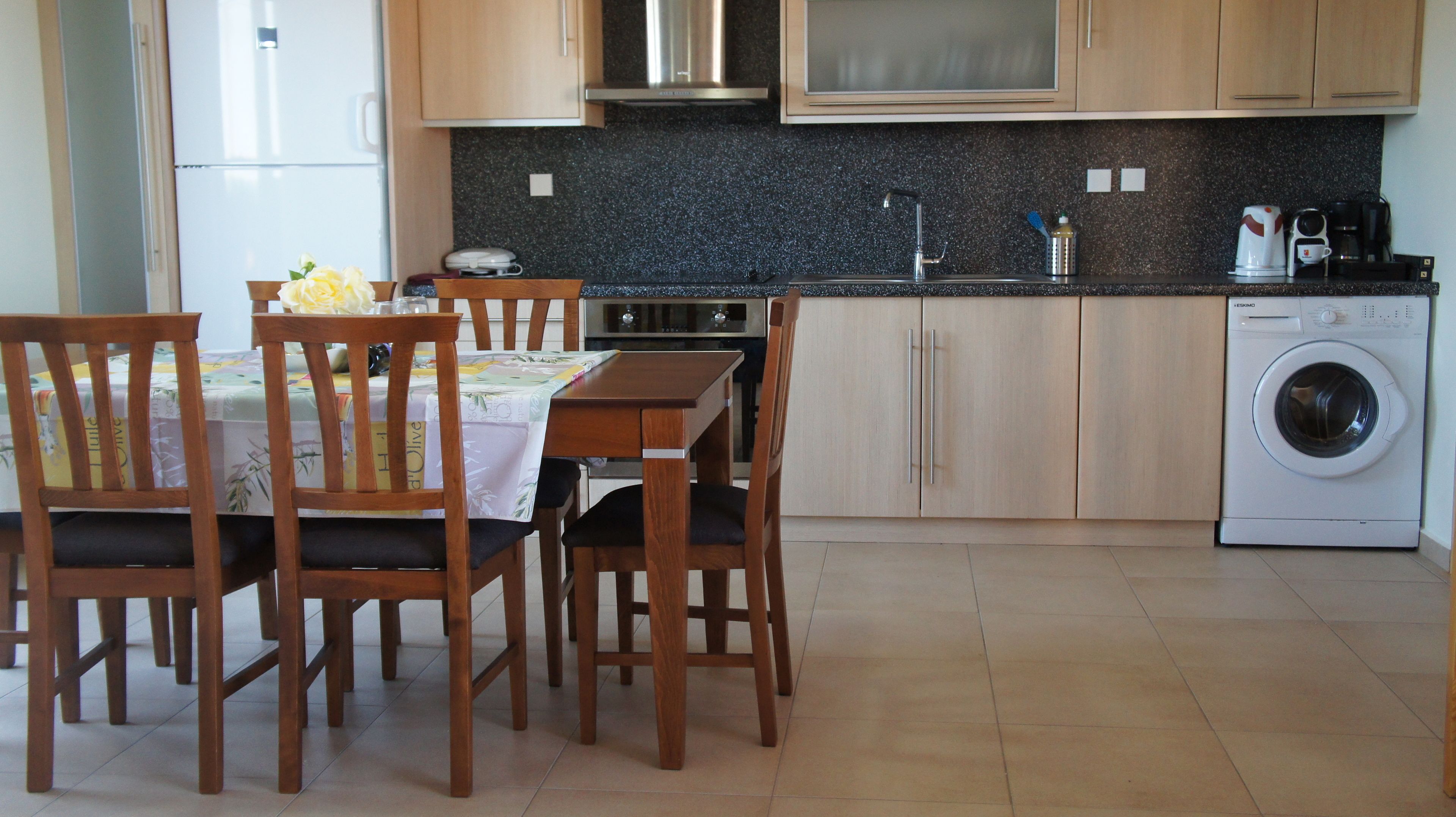 A kitchen with wooden cabinets, a dining table with chairs, a washing machine, and a coffee maker.
