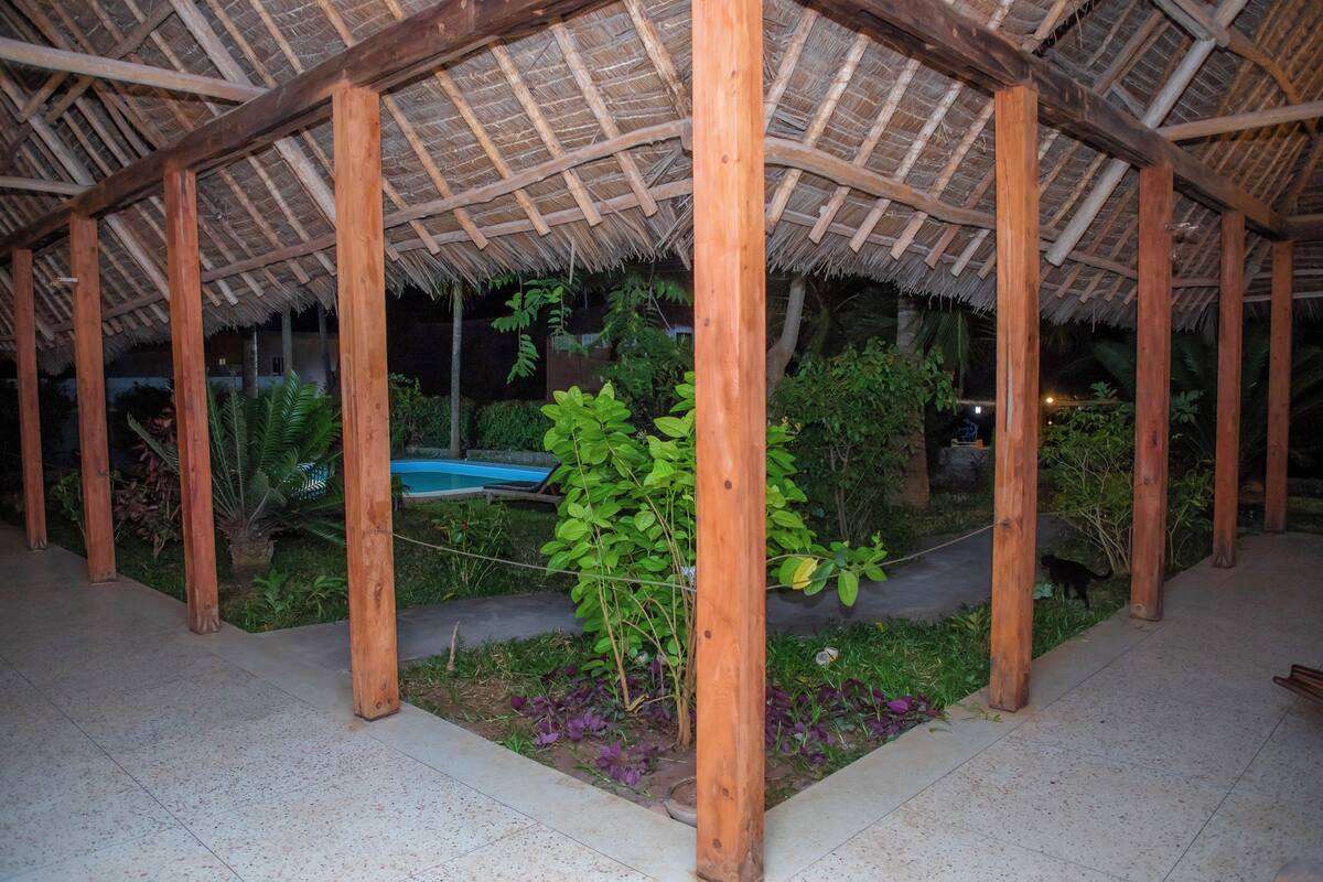 A covered outdoor area with wooden beams and a thatched roof, surrounded by greenery and a small pool.