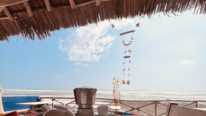 Une table installée sur la plage, recouverte d’une nappe colorée, avec une théière blanche, un pot à café en métal et une assiette remplie de fruits et de collations.