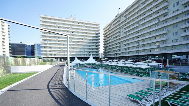 Une piscine entourée d’une clôture, dotée de chaises longues et de parasols, avec des bâtiments modernes en arrière-plan.