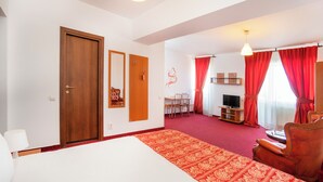 Quarto de hotel com uma cama grande, uma escrivaninha com cadeira, televisão e cortinas vermelhas.