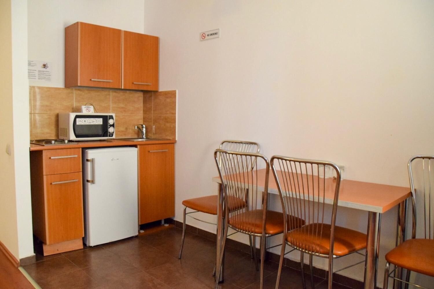 A compact kitchen with wooden cabinets, a microwave, and a small refrigerator. There is a dining table with four metal chairs.