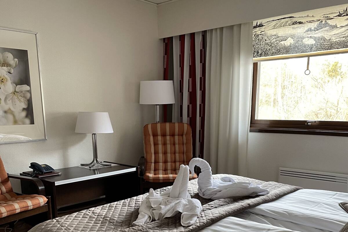 A hotel room with a bed, a chair, a desk, a lamp, and a window with a view of trees.