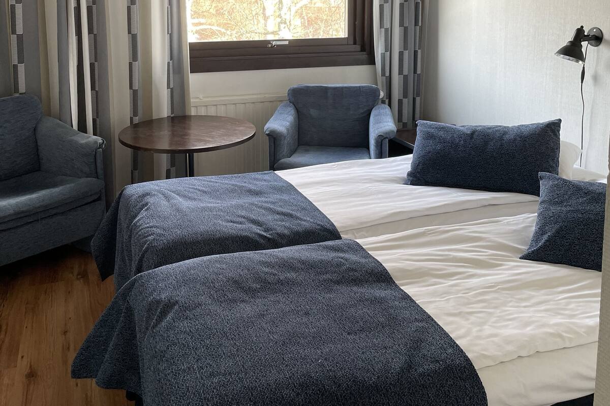 A hotel room with a large bed, two bedside tables, a sofa, and a chair. There is a window with curtains, and a lamp on the wall.