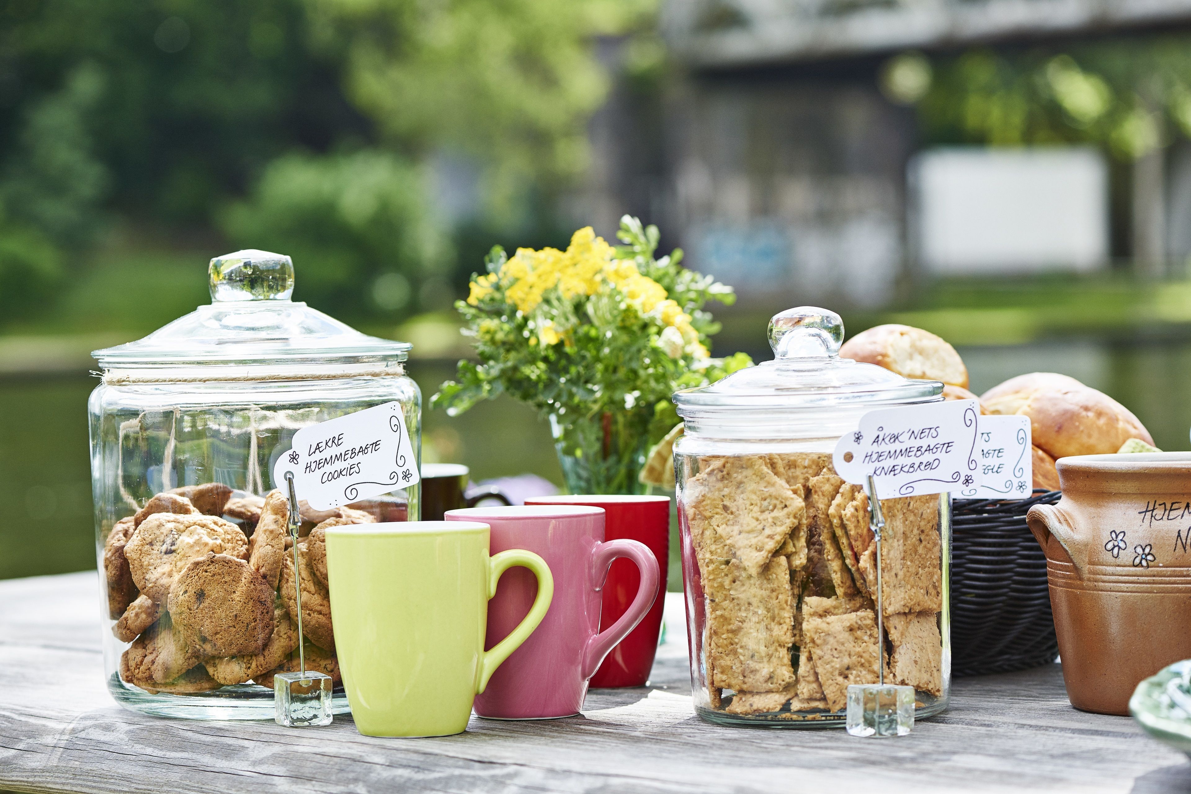 A picnic setup with jars of cookies and biscuits, colorful mugs, and a basket of bread on a wooden table by a body of water.