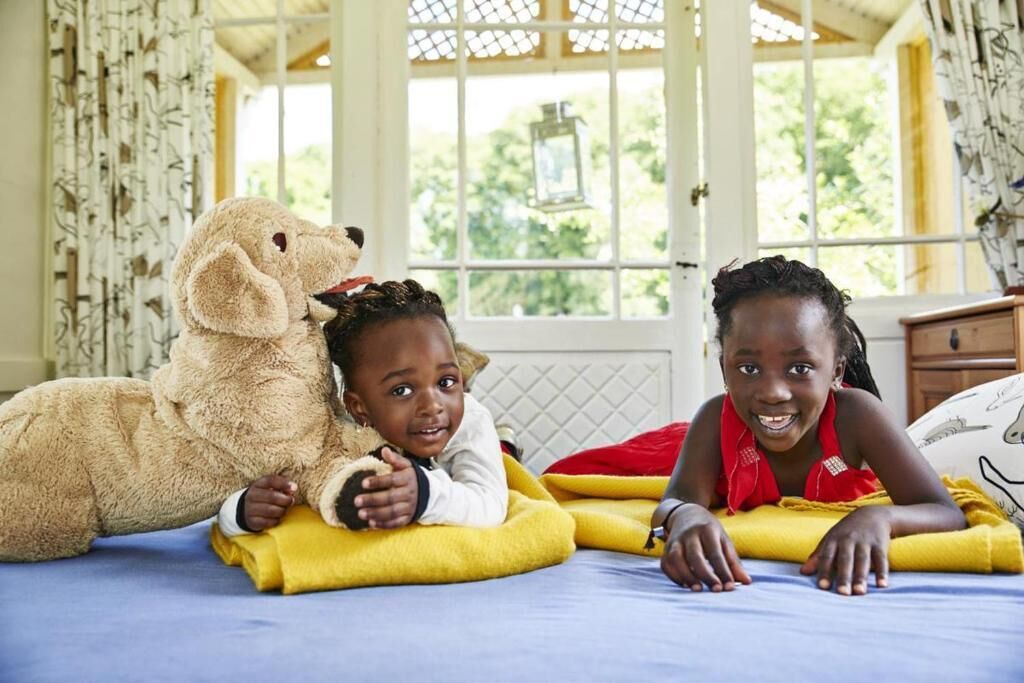Deux enfants allongés sur un lit, avec un grand ours en peluche, dans une pièce dotée d’une fenêtre et de rideaux.