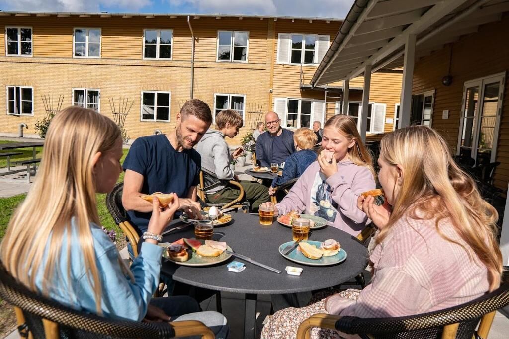 Un groupe de personnes est réuni autour d’une table ronde à l’extérieur, où elles dégustent des plats et des boissons, devant un bâtiment doté de nombreuses fenêtres.