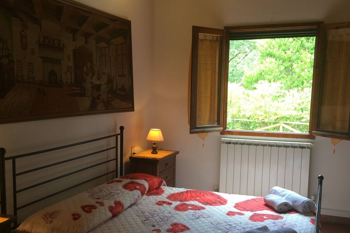 Ein Schlafzimmer mit einem Bett, einem Nachttisch, einer Lampe, einem Fenster mit Vorhängen und einem Bild an der Wand.