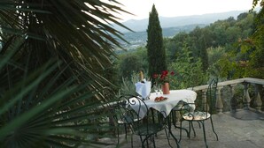 An outdoor dining area with a table set for two, surrounded by metal chairs, a stone balustrade, and a view of a mountainous landscape. - Podere San Lorenzo (Terranuova Bracciolini)