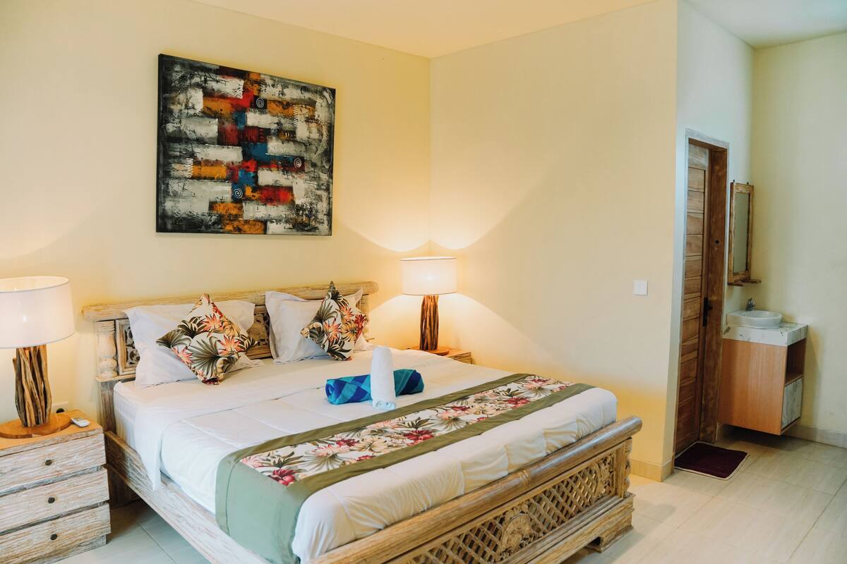 A neatly arranged bedroom with a bed, bedside lamps, a wooden nightstand, a mirror, and a bathroom with a sink and mirror.