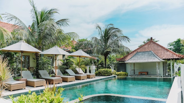 Un espace piscine agrémenté de chaises longues et de parasols, entouré d’une végétation luxuriante et bordé d’un bâtiment traditionnel doté d’un toit en tuiles.
