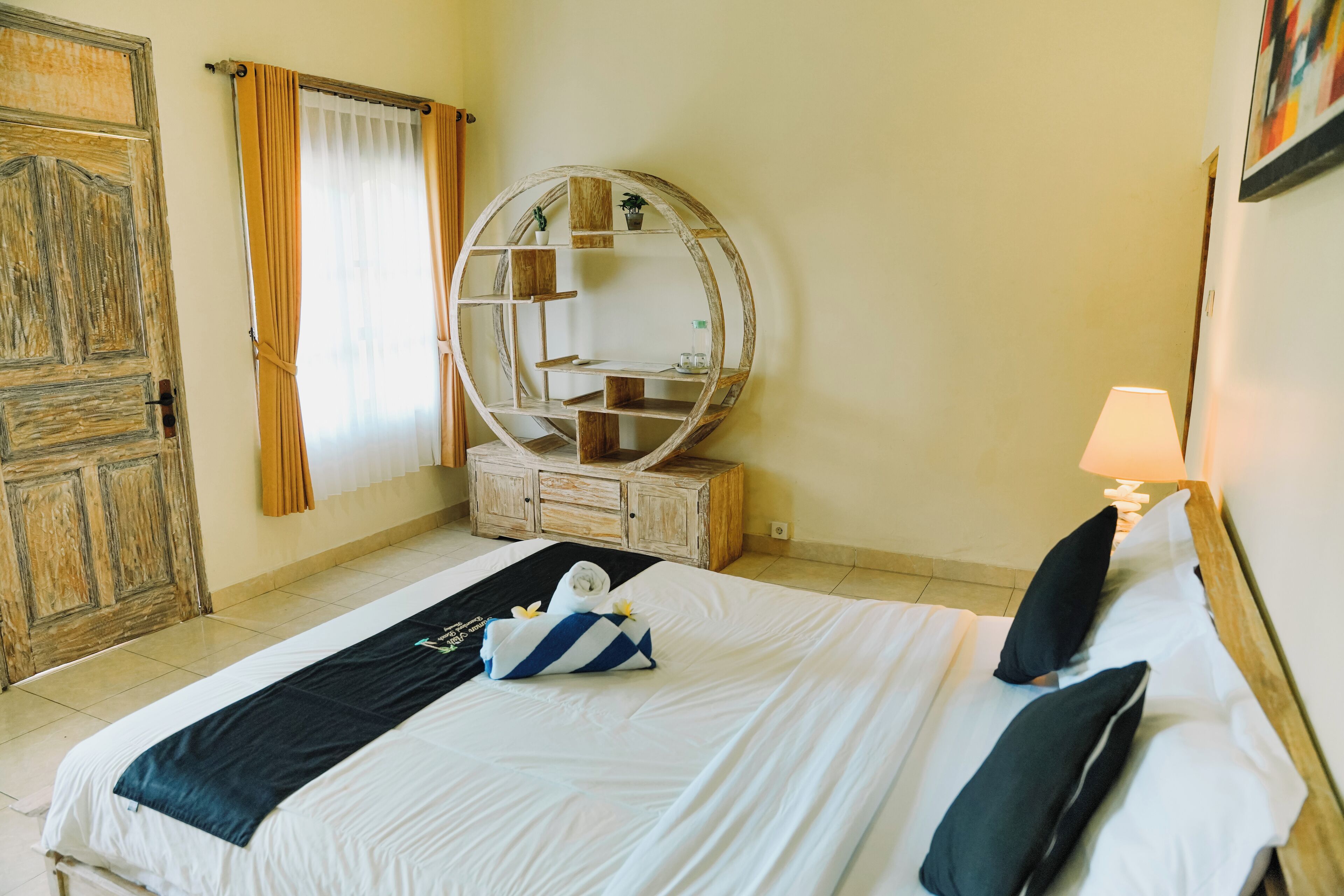 Un lit bien fait, avec une serviette à rayures bleues et blanches, une commode en bois avec un miroir et une lampe sur la table de chevet, dans une chambre d’hôtel.