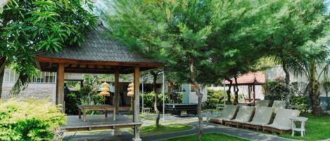 A covered outdoor seating area with a table and chairs, surrounded by lush greenery and trees.