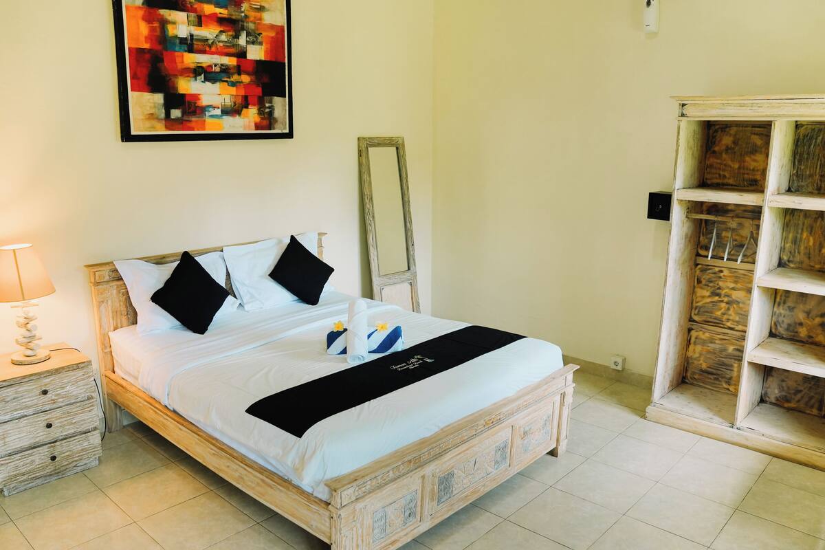 A neatly made bed with a black bedspread, two pillows, and a towel with a yellow flower. There is a wooden nightstand with a lamp and a drawer, a mirror on the wall, and a wooden wardrobe with shelves.