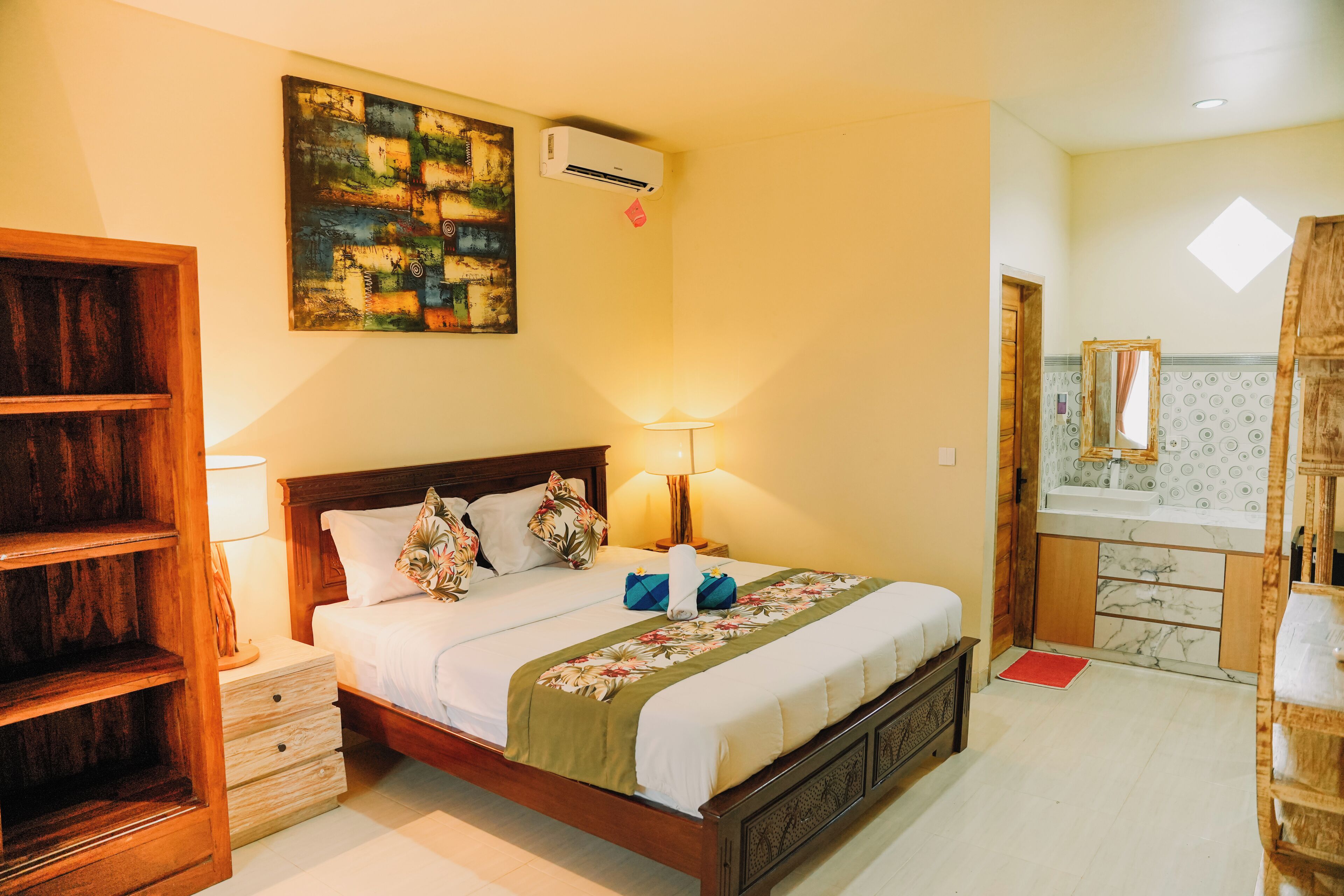 Une chambre d’hôtel avec un lit en bois, une table de chevet, une lampe, un tableau et une salle de bain visible à travers une porte ouverte.