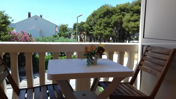 Un balcone con un piccolo tavolo e due sedie di legno, una pianta in vaso fiorita, e vista su alberi ed edifici.