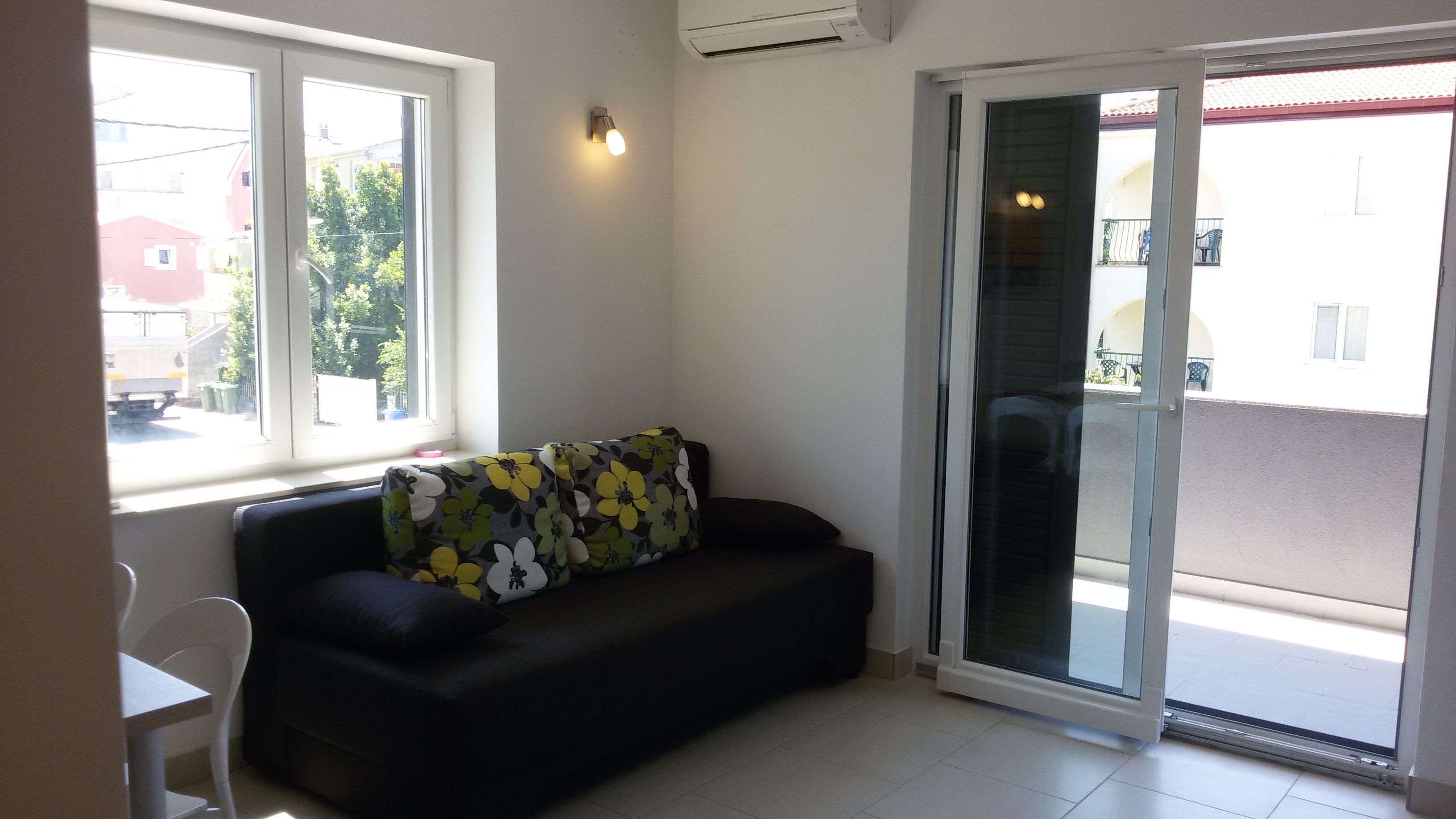 Ein Wohnzimmer mit einer schwarzen Couch, einem weißen Sessel und einem Fenster mit Blick auf eine Straße und Gebäude.