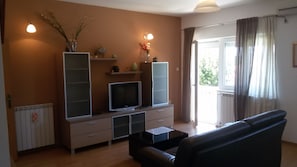Ein Wohnzimmer mit Fernseher, einer Couch, einem Couchtisch und einem Schrank. Ein Fenster mit Vorhängen und eine Heizung sind ebenfalls vorhanden.