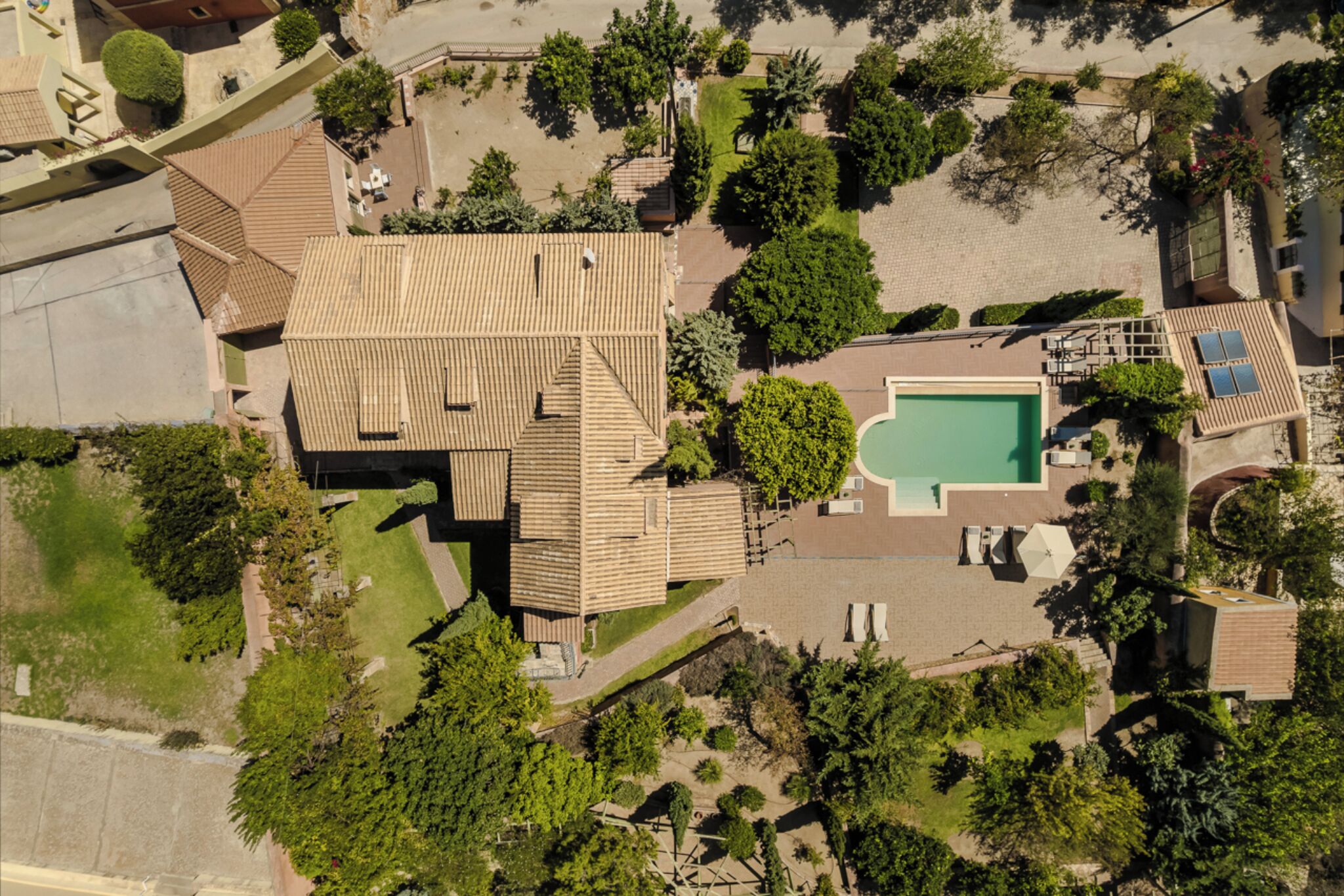 An aerial view of a residential area with a swimming pool, a large house with a tiled roof, and surrounding greenery.