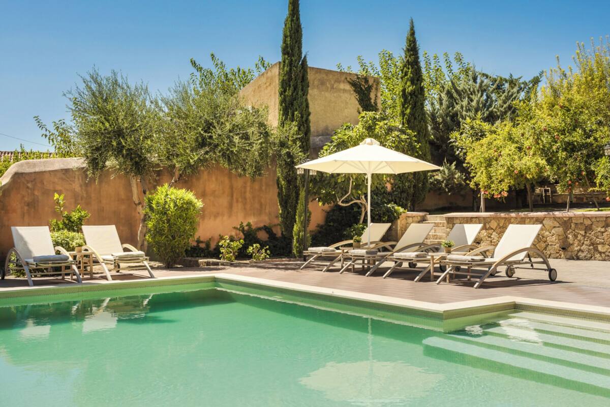 A swimming pool with lounge chairs and an umbrella, surrounded by greenery and a stone wall.