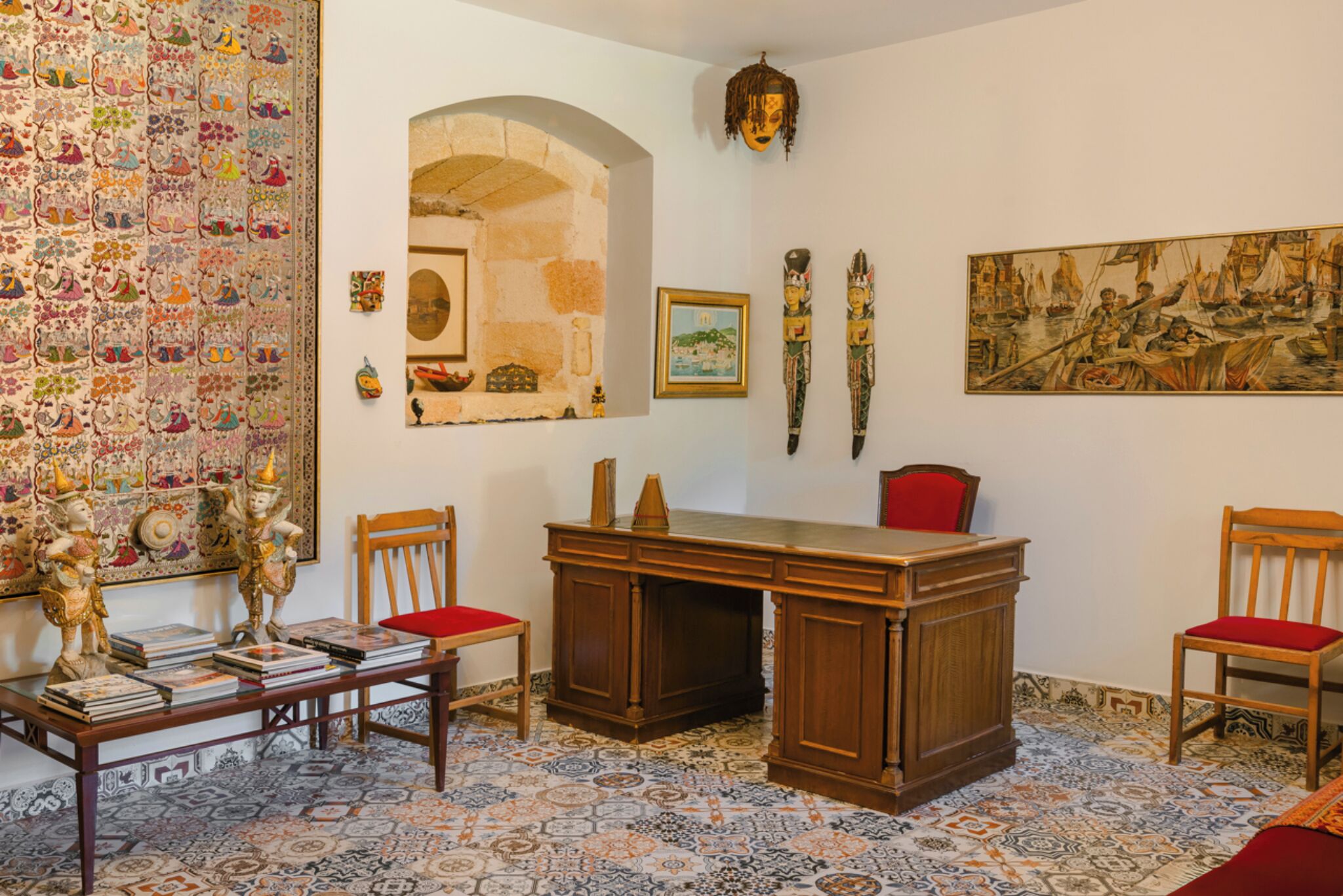 A room with a wooden desk, chairs, a patterned carpet, a tapestry, and wall-mounted artwork.